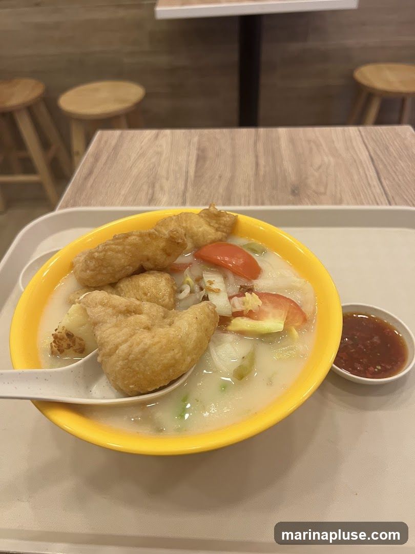 A close-up shot of a yellow bowl filled with a creamy soup. The soup contains chunks of white fish, sliced tomatoes, and green vegetables. Several pieces of golden-brown fried fish are placed on top of the soup. A white spoon rests on the edge of the bowl, scooping up some of the soup and fish. To the right of the bowl is a small white dish containing a reddish dipping sauce. The bowl sits on a white tray, which is placed on a light-colored surface. In the background, a wooden table and two wooden stools are visible, suggesting a casual dining setting.