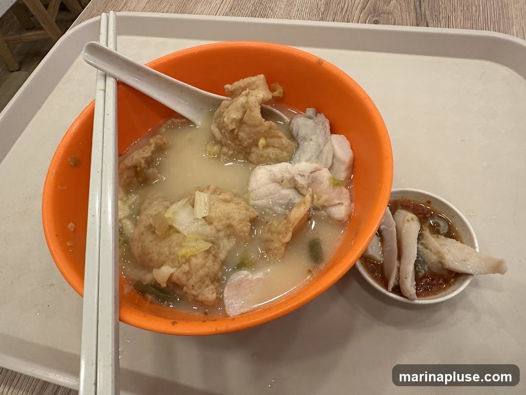 A close-up shot of a bowl of fish soup served on a white tray. The soup contains fish balls, chunks of fish, and some vegetables. A small dish of dipping sauce is placed next to the main bowl. Chopsticks and a spoon rest on the rim of the bowl.