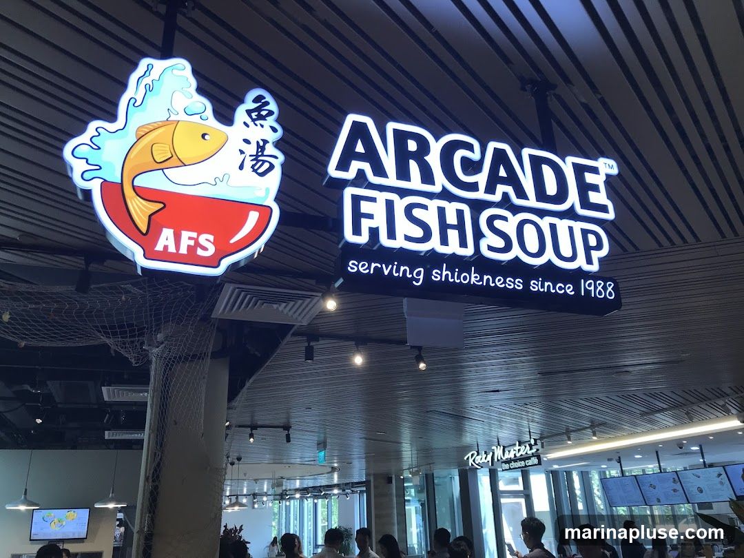 A brightly lit sign for 'Arcade Fish Soup' hangs from the ceiling, featuring a cartoon fish leaping out of a bowl. Below the main text, it reads 'serving shiokness since 1988'. In the background, a food court setting is visible with other establishments and patrons.