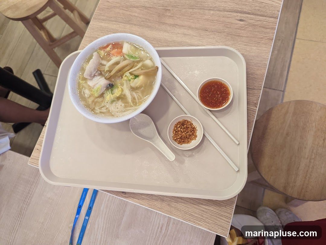 A bowl of noodle soup with fish, vegetables, and mushrooms is served on a tray with chopsticks, a spoon, a small bowl of chili sauce, and a small bowl of fried shallots. The tray is placed on a wooden table.