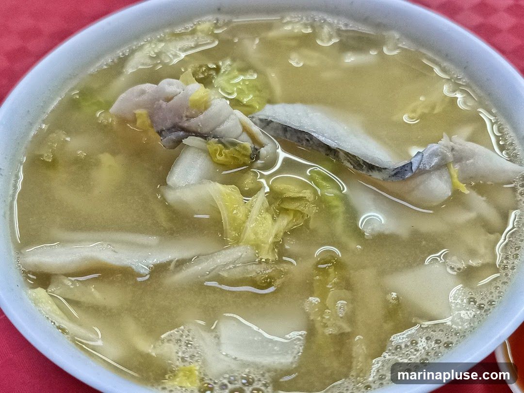 A close-up shot of a bowl of fish soup with chunks of fish and leafy vegetables. The broth is a light yellow color and appears to have some oil on the surface. The fish pieces are white and flaky, with some skin still attached. The vegetables are green and wilted. The bowl is light blue and sits on a red tablecloth.