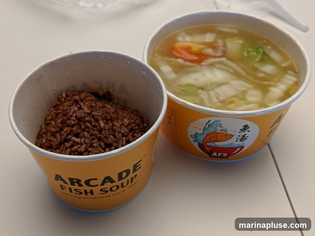 A close-up shot of two paper containers, one filled with brown rice and the other with fish soup. The container with rice has the text "ARCADE FISH SOUP serving shiokness since 1988" printed on it. The soup container has a logo with a fish and the text "AFS 魚湯" (AFS Fish Soup). The soup contains cabbage and tomato slices.