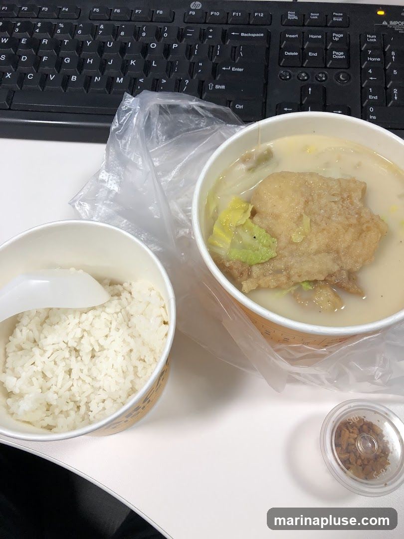 A meal consisting of a bowl of white rice and a bowl of soup with a piece of fried food and vegetables. A small container of fried garlic bits is also visible. The meal is placed on a white surface next to a black computer keyboard.