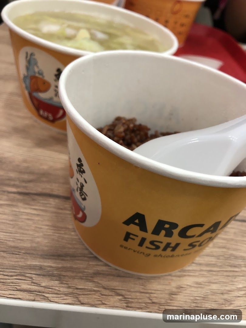 Two paper cups with branding for "Arca Fish Soup" are on a wooden table. One cup contains a light-colored soup with vegetables, and the other contains a portion of brown rice with a white spoon.