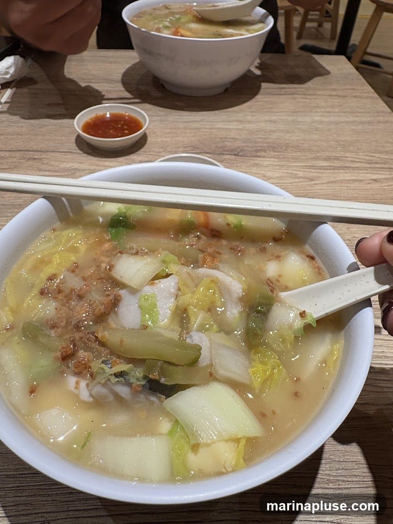 A close-up view of a bowl of fish soup with vegetables and a side of chili sauce. The soup contains pieces of fish, napa cabbage, and other vegetables. Chopsticks are resting on the rim of the bowl. Another bowl of soup is visible in the background.