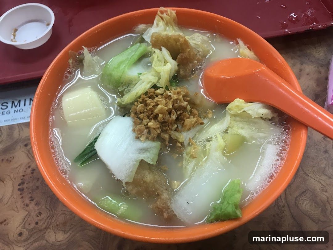 A close-up shot of a bowl of soup filled with various ingredients. The soup contains chunks of white vegetables, possibly cabbage or bok choy, and some pieces of fried batter. It is topped with a generous amount of crispy fried garlic. An orange spoon rests on the side of the bowl. In the background, a small white dish with some remnants of food and a red tray are visible. A portion of a printed document with text is also visible in the lower left corner.