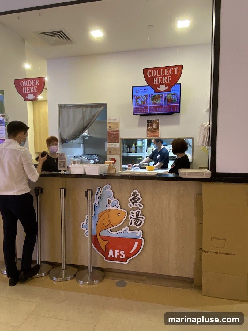 A view of a restaurant counter with staff serving customers. There are signs indicating "ORDER HERE" and "COLLECT HERE". A digital menu is displayed on a screen above the counter. The counter itself has a logo featuring a fish in a bowl with the text "AFS" and Chinese characters. A cardboard box labeled "5oz Food Container" is visible to the right.