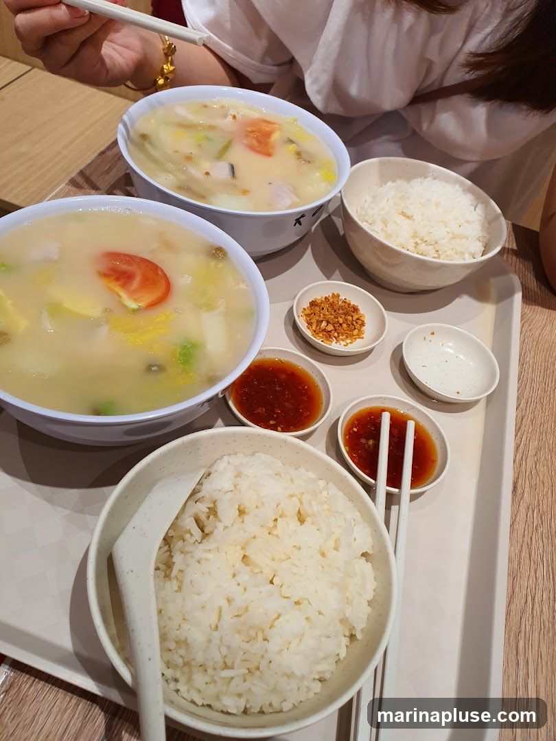 A tray holds two bowls of soup with vegetables and meat, two bowls of white rice, and small dishes of chili sauce and fried garlic. Chopsticks are visible.