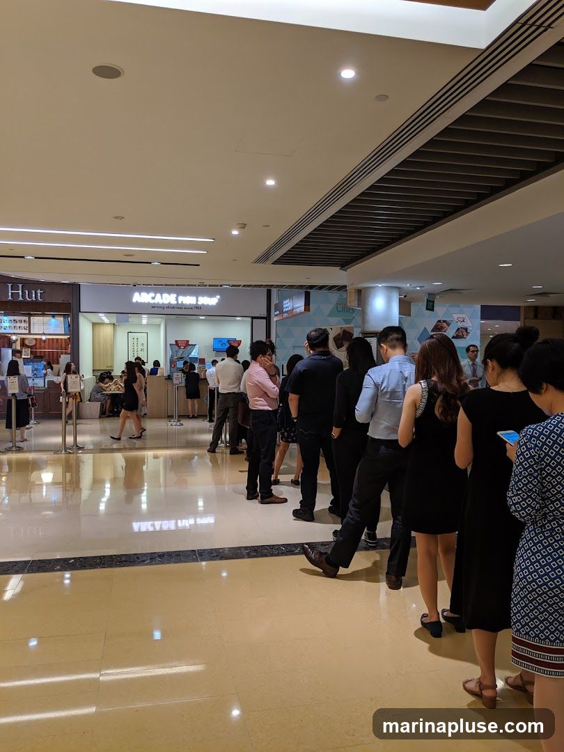 A long queue of people are waiting to be served at a food stall in a mall. The stall has a sign that says "ARCADE FISH SOUP" and "serving deliciousness since 1988". There are other shops visible in the background, including one with a sign that says "Hut". The floor is highly reflective, showing the lights and ceiling of the mall.