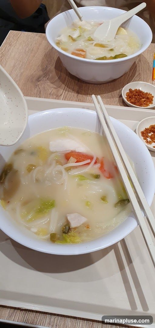A close-up view of two bowls of noodle soup served on a tray. The soup contains noodles, chopped vegetables such as cabbage and carrots, and pieces of meat or fish. Chopsticks and spoons are placed in the bowls. Small dishes of fried shallots are on the side. The table has a wood-grain pattern.