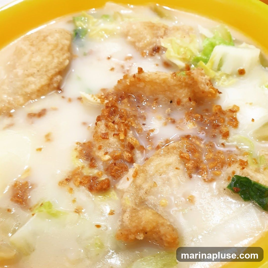 A close-up shot of a creamy soup served in a yellow bowl. The soup contains pieces of fried food, possibly fish or tofu, and chopped vegetables like napa cabbage. Crispy fried garlic bits are sprinkled on top of the fried pieces.