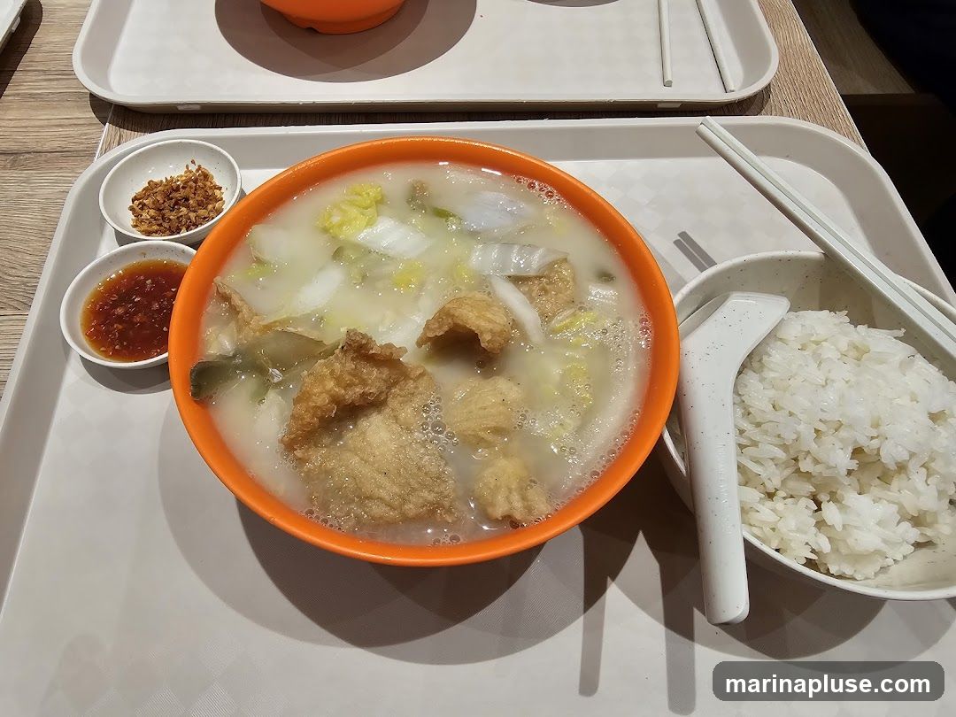 A bowl of fish soup with vegetables and fried fish pieces, served with a side of rice, chili sauce, and fried shallots. The meal is presented on a tray with chopsticks and a spoon.