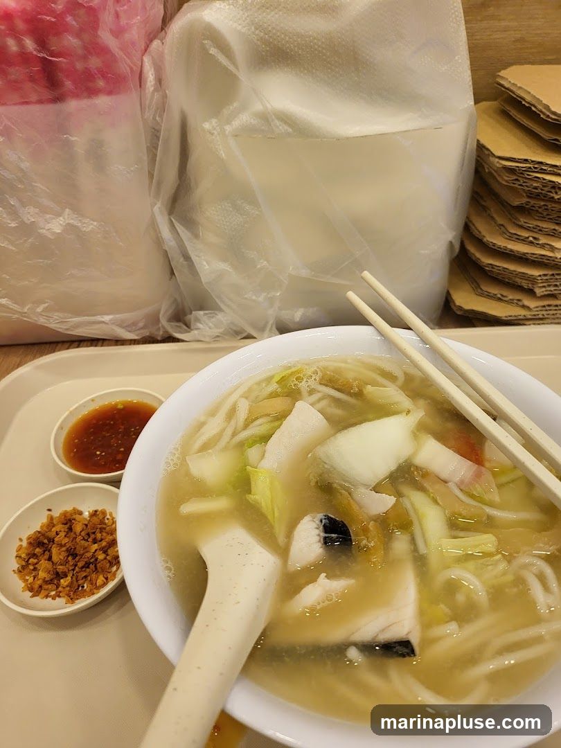 A bowl of fish soup noodles with a spoon and chopsticks. Two small dishes of condiments, one with chili sauce and the other with fried garlic, are placed to the left of the bowl. In the background, there are plastic bags and stacks of cardboard.