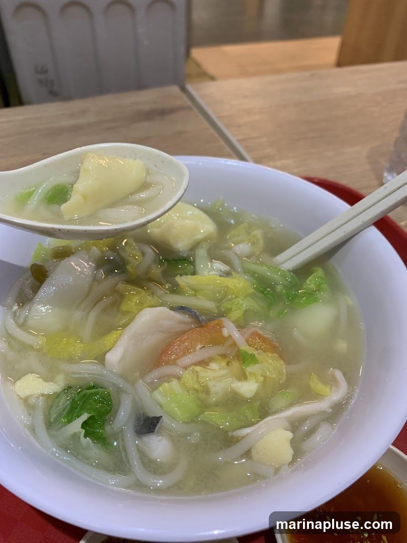 A close-up shot of a bowl of fish soup with noodles, vegetables, and pieces of fish. A spoon is lifting some of the soup and ingredients, showing tofu, cabbage, and noodles. The bowl is on a red tray, and a small dish of dipping sauce is visible at the bottom right.
