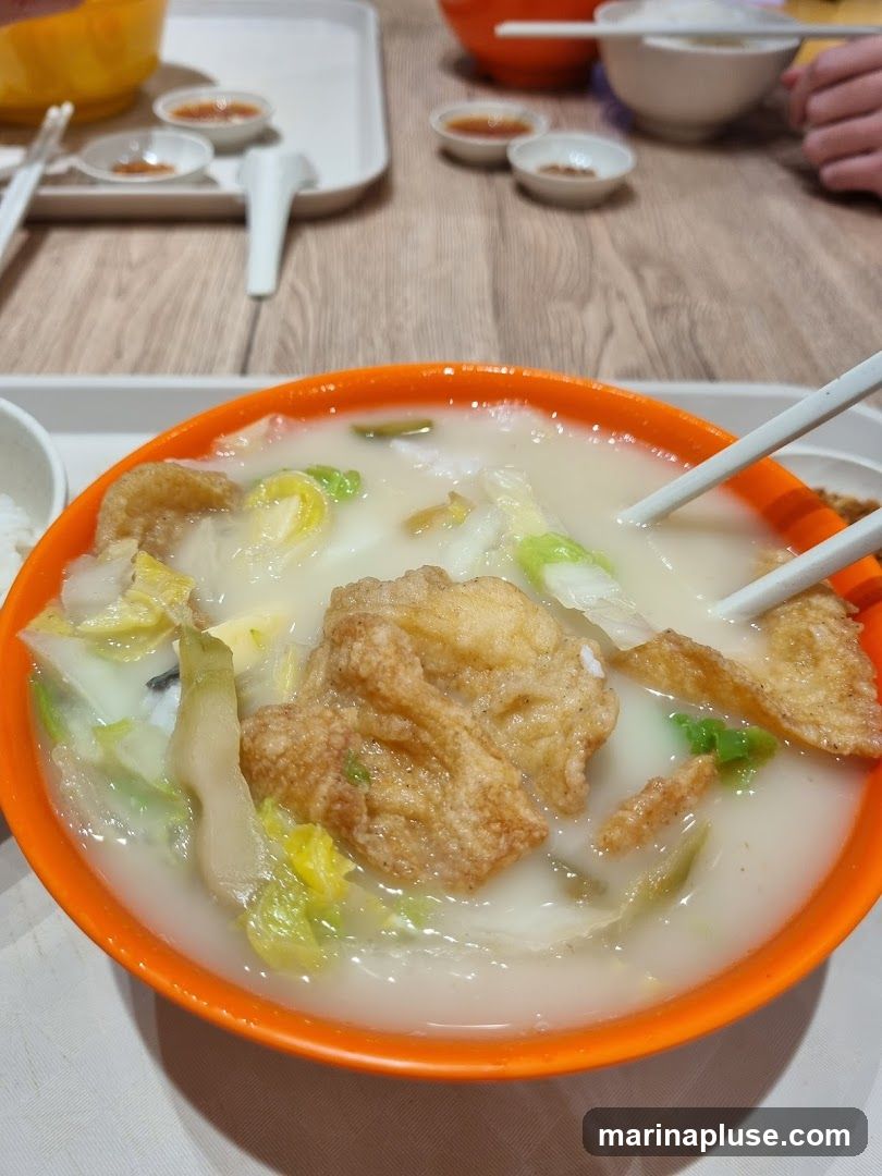A close-up shot of a bowl of fish soup with vegetables and fried fish pieces. The soup is milky white and contains chunks of cabbage and other greens. Chopsticks are visible, holding a piece of fried fish. In the background, there are small dishes with dipping sauces and bowls of rice.