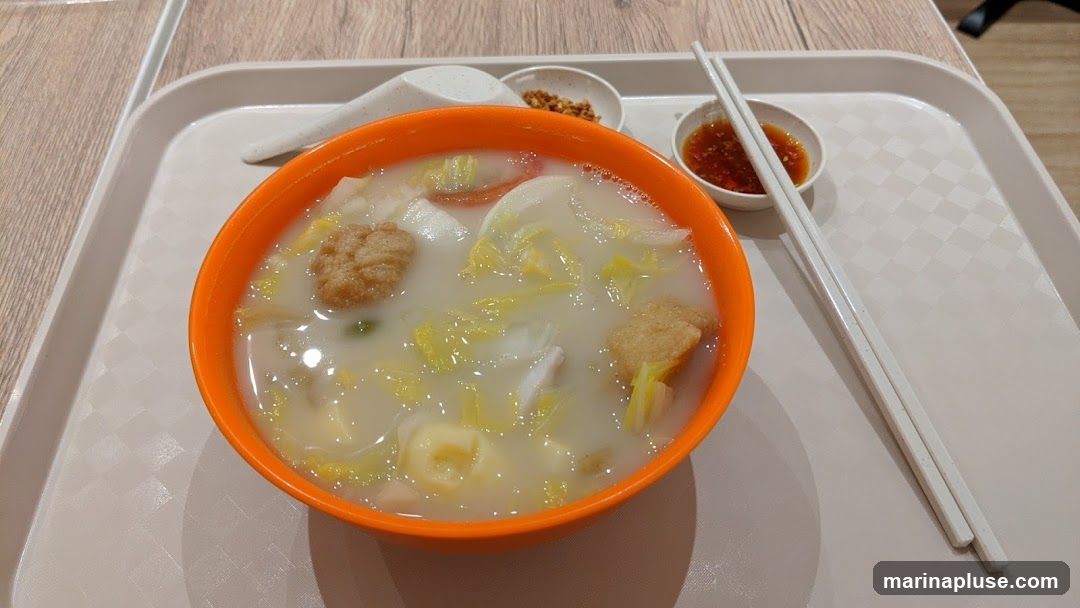 A close-up shot of a bowl of soup with noodles, vegetables, and fried tofu. The soup is served in an orange bowl and placed on a white tray with a spoon and chopsticks. There are two small dipping sauces on the side.