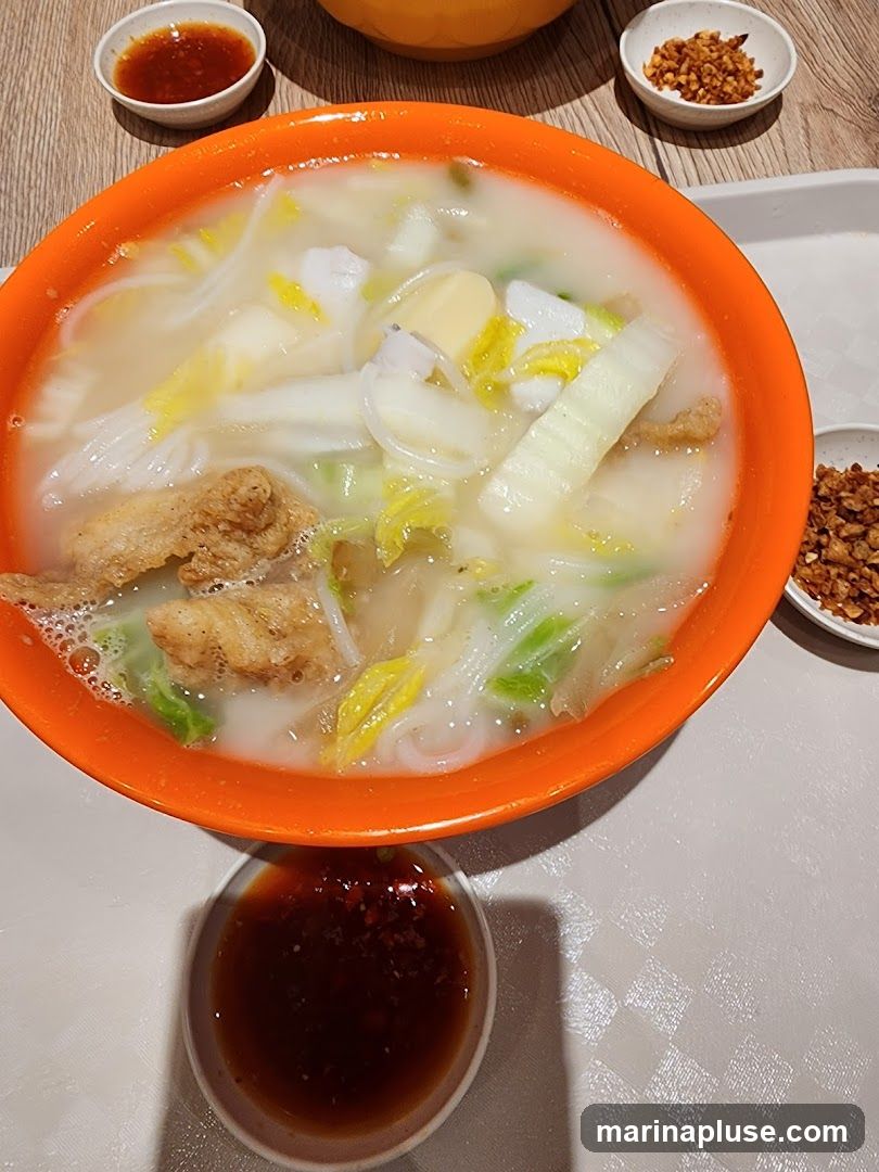 A close-up overhead view of a bowl of soup with noodles, fish, tofu, and leafy green vegetables. There are three small dipping bowls surrounding the main bowl: one with a dark sauce, one with chili flakes, and another with a reddish sauce. The soup is served in an orange bowl.