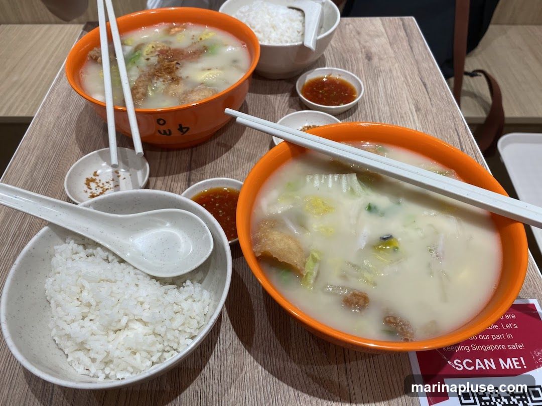 Two orange bowls of soup with various ingredients and white chopsticks are placed on a wooden table. Next to the bowls are smaller dishes with chili sauce and what appears to be fried garlic or shallots. Two bowls of white rice are also visible, one with a spoon resting on it. A red and white sign with a QR code and the text "SCAN ME!" is on the table.