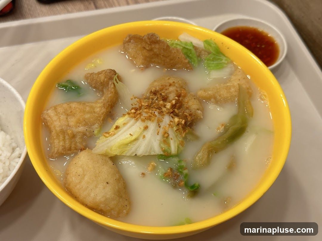 A close-up shot of a bowl of fish soup served with rice. The soup contains fried fish pieces, napa cabbage, and other vegetables, topped with fried garlic. A small dish of chili sauce is on the side.
