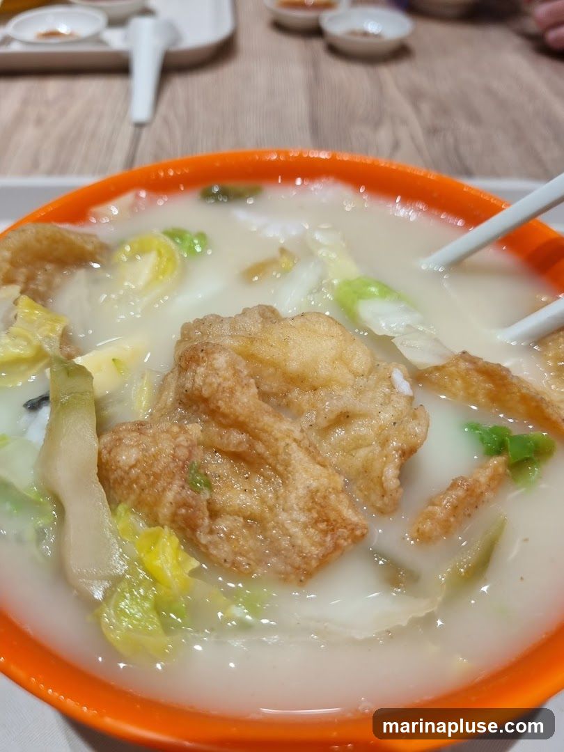 A close-up shot of a bowl of fish soup with fried fish pieces, cabbage, and other vegetables. The soup is milky white and served in an orange bowl. Chopsticks are visible in the soup. In the background, there are small dipping bowls and a white tray.