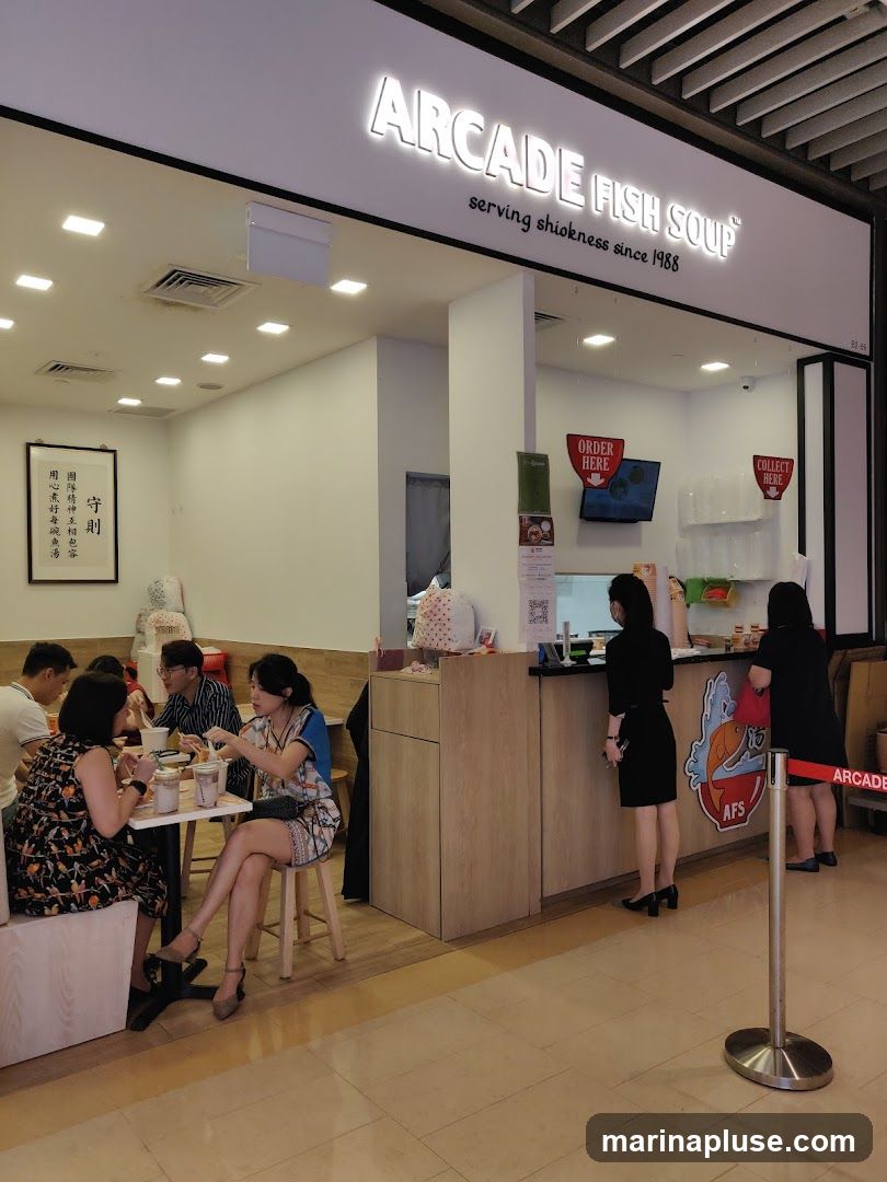 A brightly lit storefront for Arcade Fish Soup, with a large illuminated sign above the counter. The sign reads "ARCADE FISH SOUP" and "serving shiokness since 1988". Inside, customers are seated at tables, eating. Staff members are visible behind the counter. There are signs for "ORDER HERE" and "COLLECT HERE".