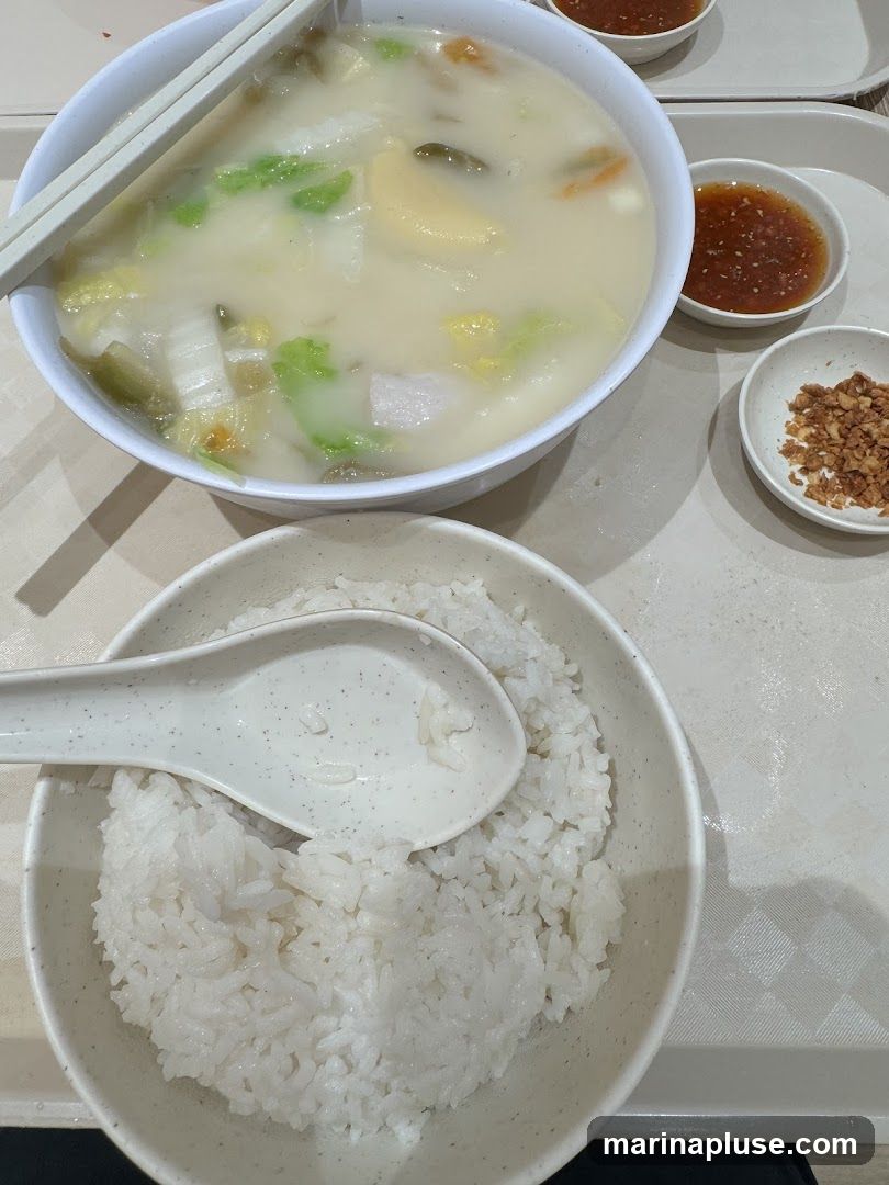 A bowl of soup with vegetables and meat, served with a bowl of rice, a spoon, chopsticks, and two small dipping sauces. One sauce is red and appears to be chili-based, while the other is a pile of fried garlic bits.
