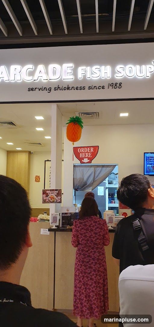 The exterior of a fish soup restaurant with a prominent sign that reads "ARCADE FISH SOUP serving shiokness since 1988". An orange pineapple-shaped decoration hangs above the counter, and a red "ORDER HERE" sign is visible. People are queuing at the counter.
