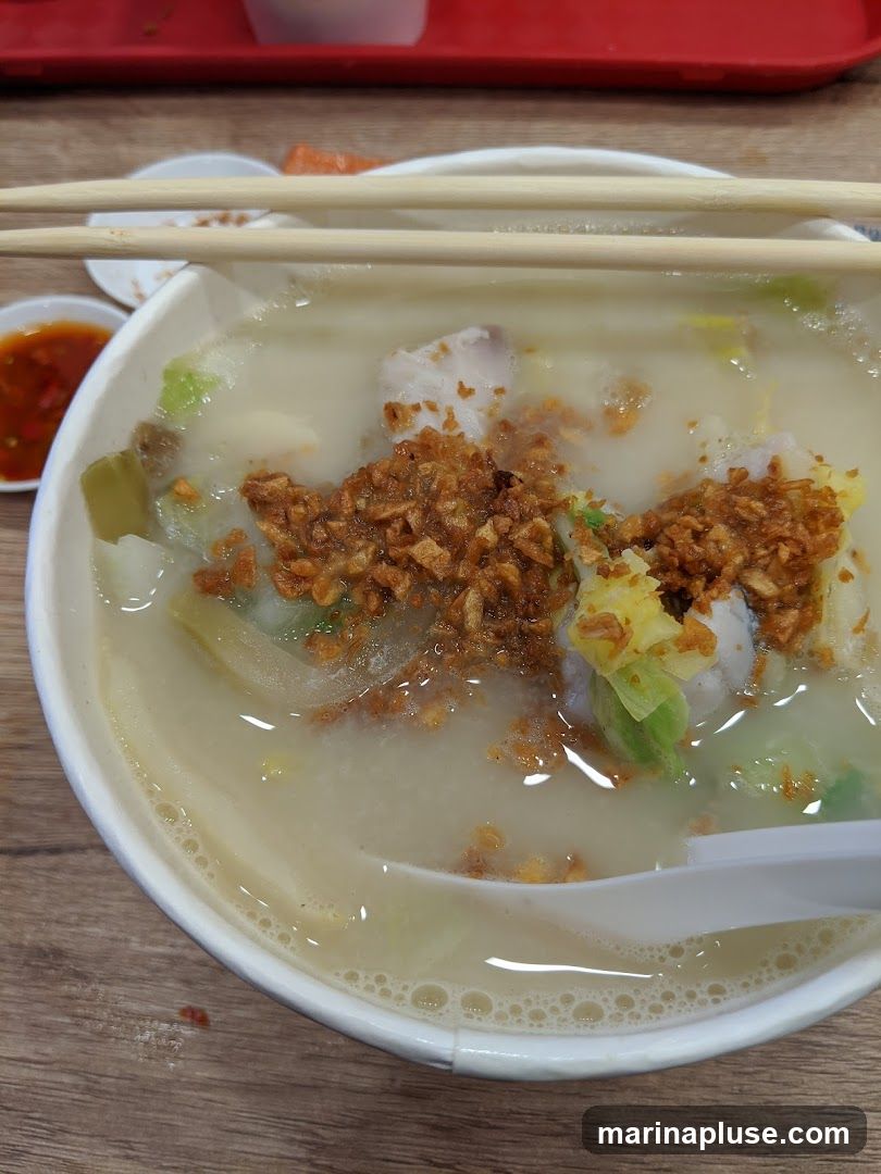A close-up shot of a bowl of fish soup, topped with fried garlic and pieces of fish and vegetables. Chopsticks rest on the rim of the bowl, and a small dish of chili sauce is visible to the left. The soup appears milky and is served in a white disposable bowl.