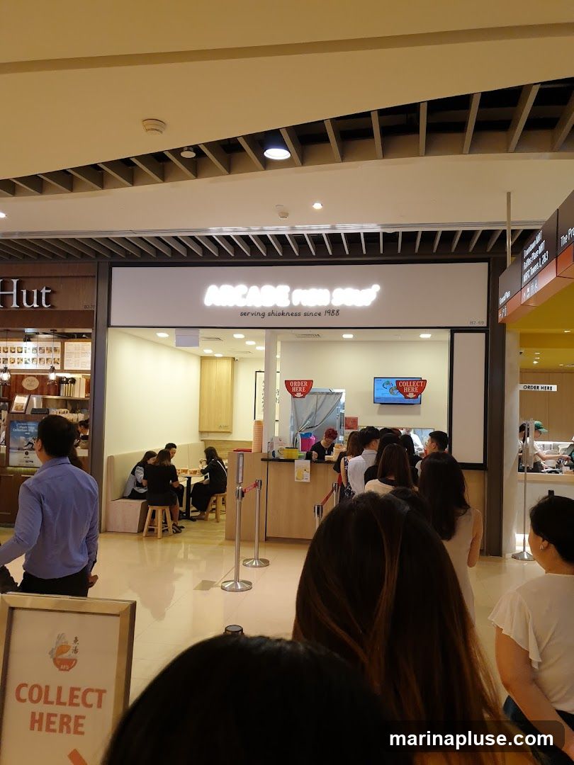 A queue of people waiting to order at a food stall called Arcade Fish Head Noodles, which has been serving since 1988. The stall is located in a mall, with other shops visible in the background. There are signs indicating 'Order Here' and 'Collect Here'.