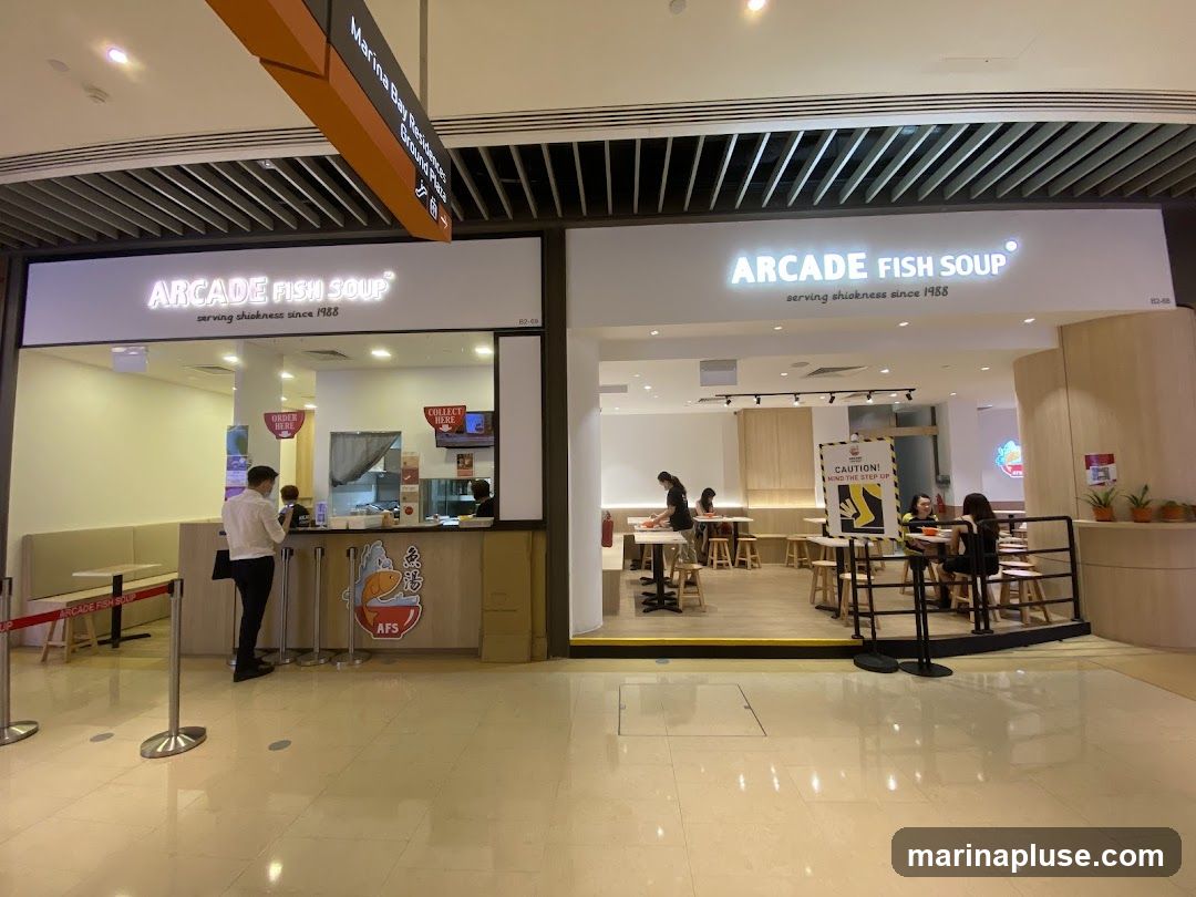 The exterior of two adjacent Arcade Fish Soup storefronts. The left storefront has a counter with staff and customers, and seating. The right storefront has tables and chairs for dining, and a caution sign. Both storefronts have the Arcade Fish Soup logo and tagline visible.