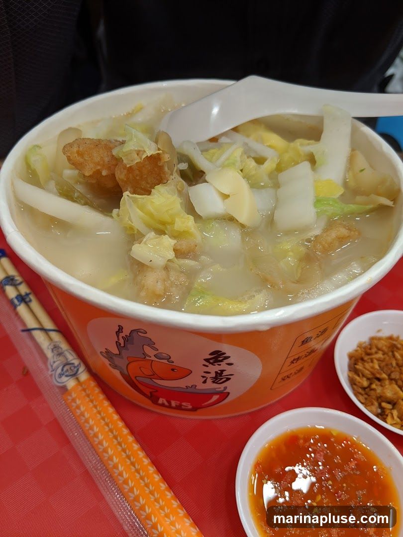 A close-up of a bowl of fish soup with vegetables and fried fish pieces. The bowl has an orange label with a cartoon fish and Chinese characters. Chopsticks in a plastic wrapper and two small dipping sauces are also visible.