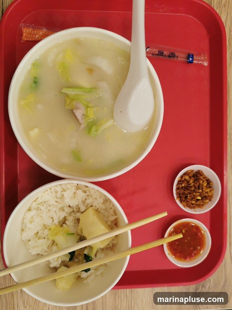 A bowl of soup with cabbage and meat, a bowl of rice with tofu and vegetables, and two small bowls of condiments (fried shallots and chili sauce) are arranged on a red tray. Chopsticks and a spoon are provided.