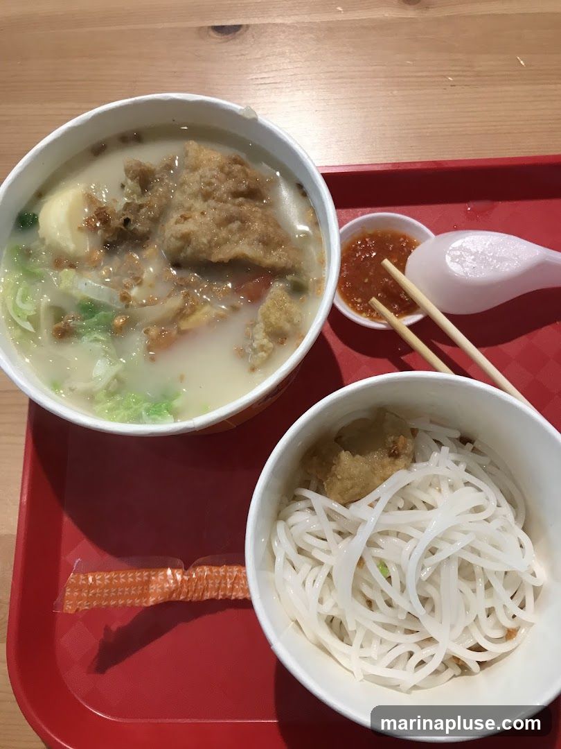 A close-up overhead shot of a meal served on a red tray. The meal consists of two white bowls, one containing noodles and the other a soup with pieces of fried fish, vegetables, and tofu. A small dish of chili sauce, chopsticks, and a spoon are also visible.