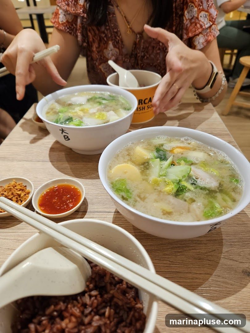 Two bowls of soup, one with tofu, vegetables, and meat, and another with similar ingredients, are placed on a wooden table. A bowl of brown rice, chopsticks, a spoon, and two small dishes of condiments (one with fried garlic and the other with chili sauce) are also visible. The background shows people dining, with a partially visible orange container that has text on it.