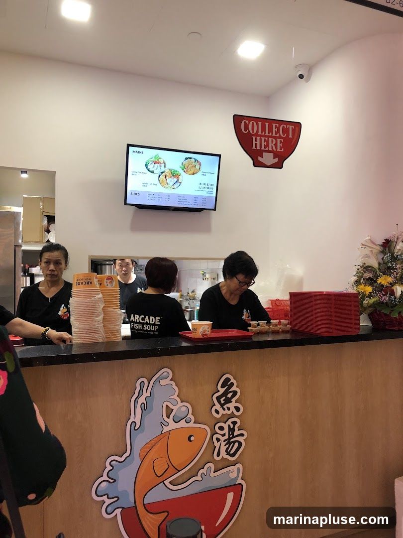 A digital menu board displays food options above a counter where staff are serving customers. A sign reads "COLLECT HERE". The counter has a wooden front with a graphic of a fish and Chinese characters. Stacks of red trays and cups are visible.