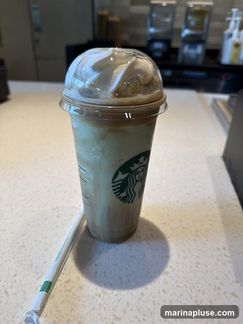 A close-up shot of a Starbucks Frappuccino with whipped cream and caramel drizzle on top. The iconic Starbucks logo is visible on the cup. A straw is placed next to the cup on a white speckled counter. In the background, there are dispensers for various liquids and a blurred counter area.