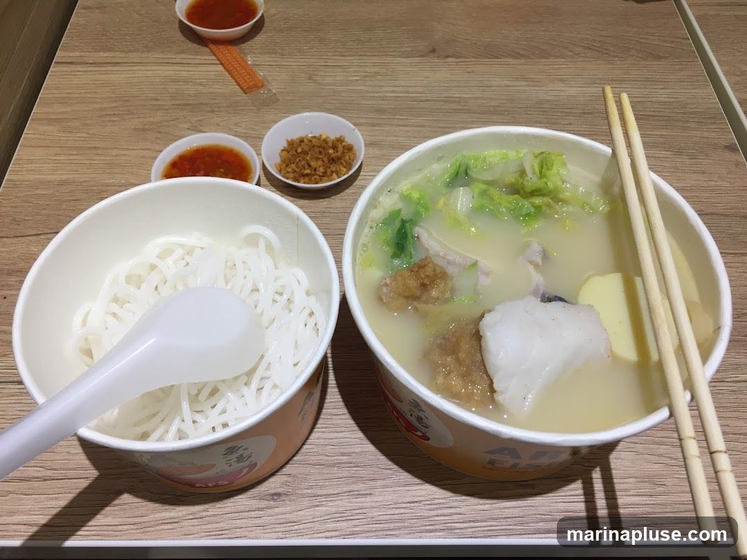 A bowl of fish soup with cabbage, tofu, and fish pieces, served with a separate bowl of noodles and a spoon. There are also two small bowls of dipping sauce and a packet of fried shallots on the side. Chopsticks are resting on the bowl of soup. The bowls have branding visible.