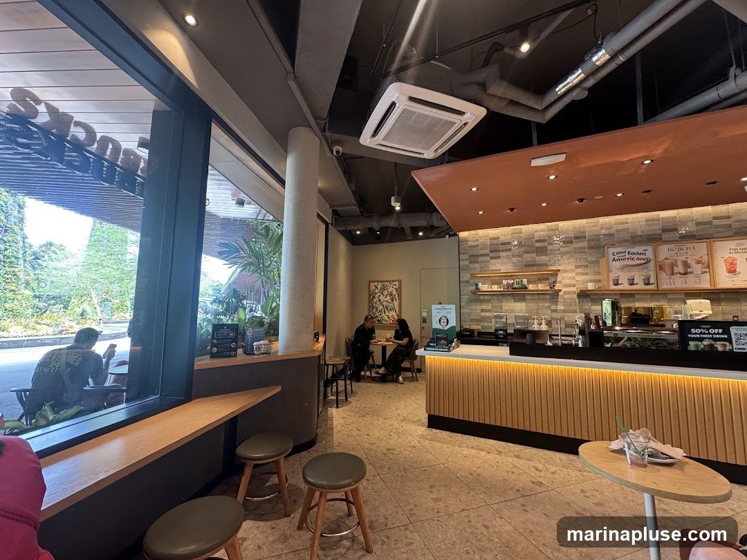 A view of the interior of a Starbucks coffee shop, looking towards the counter. The large Starbucks logo is visible through the window on the left. The counter area features menu boards displaying drinks like "Cold Foam Americano" and "Hojicha". There are also promotional signs, including one offering "50% OFF YOUR FIRST DRINK". Several customers are seated at tables and chairs, and some are ordering at the counter. The overall ambiance is modern and casual.