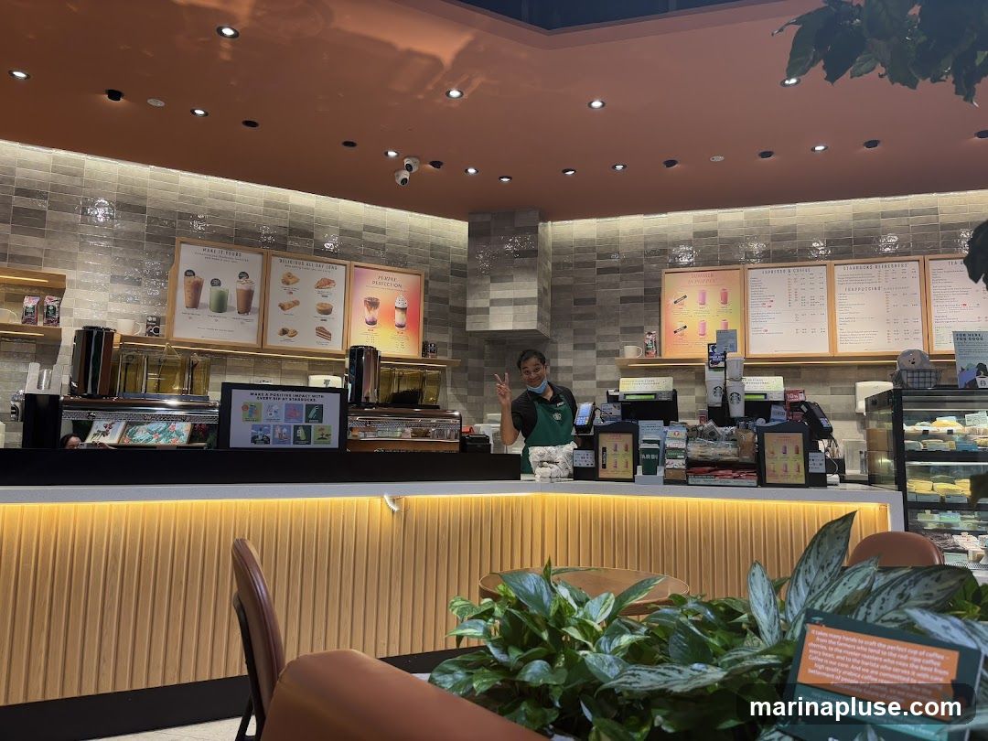 A view of a Starbucks counter with a barista smiling and giving a peace sign. There are menus displayed on the wall behind the counter, showing various drinks and food items. The counter itself has coffee machines, displays, and a refrigerated case with pastries. In the foreground, there are plants and seating areas.