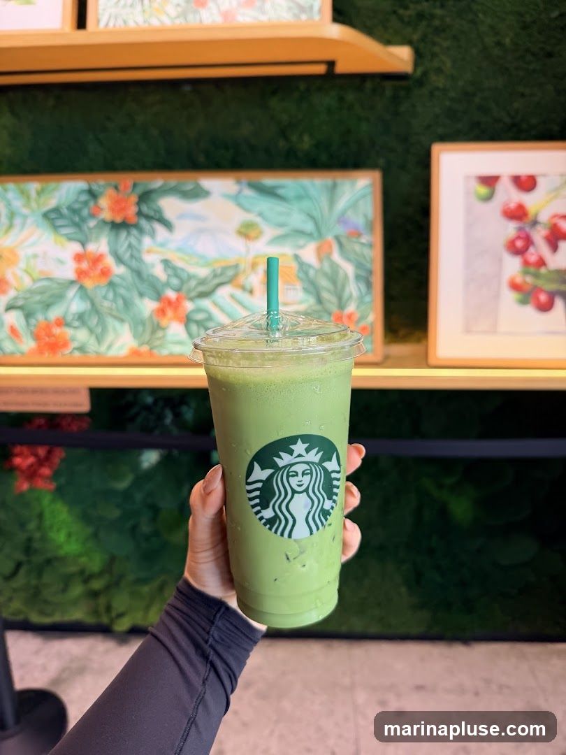 A hand holds a Starbucks matcha drink in a clear plastic cup with the Starbucks logo visible. The drink is green and appears to be iced. The background features green moss-like texture on the wall with framed artwork of tropical plants and cherries.
