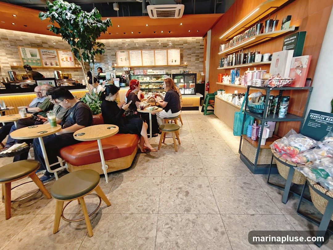 The interior of a Starbucks cafe with customers seated at tables and a counter area. Shelves display merchandise like mugs and tumblers.