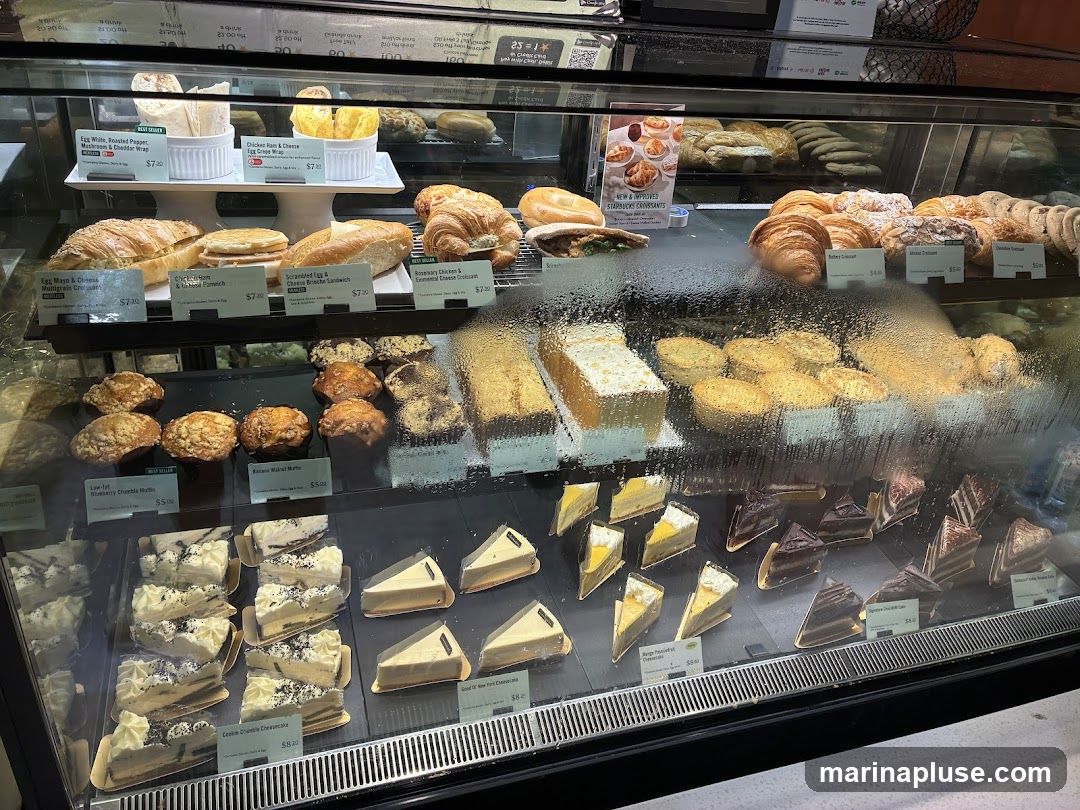 A refrigerated display case filled with various pastries, cakes, and sandwiches. The top shelf shows wraps and sandwiches. The middle shelf has croissants, danishes, and cookies. The bottom shelf displays muffins, cakes, and cheesecakes. Some items have small labels with names and prices.