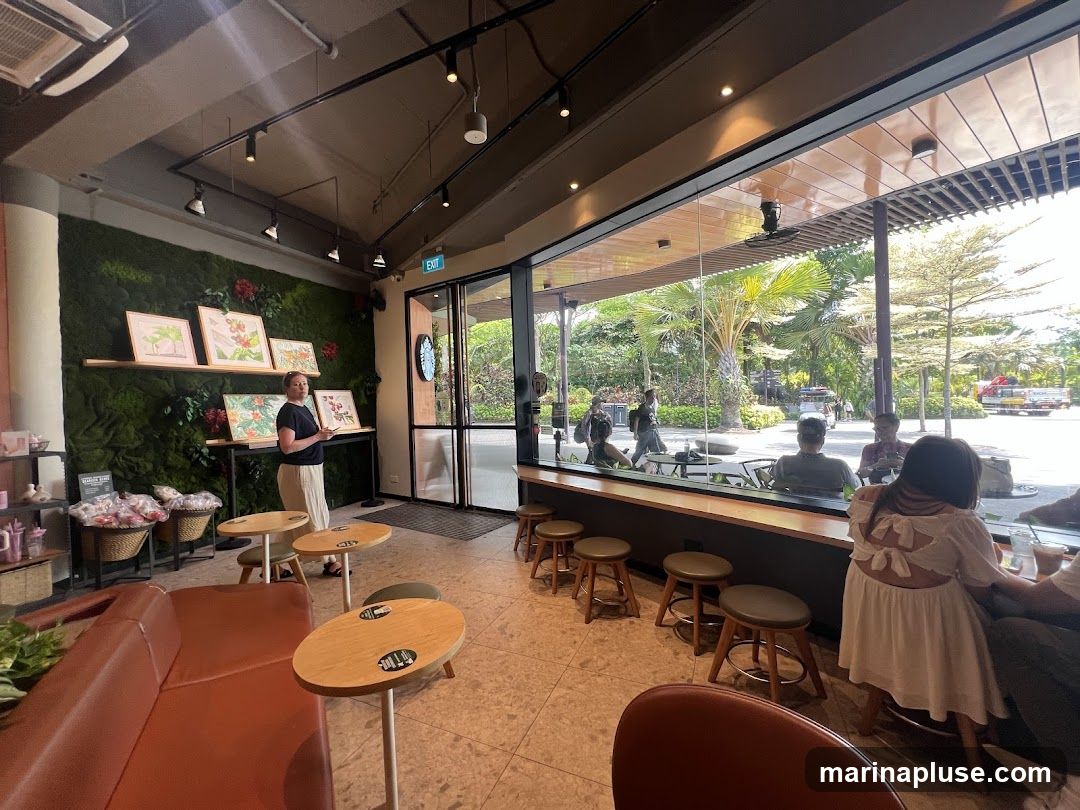 A woman stands near a green moss wall adorned with framed artwork in a cafe. The cafe has a large window overlooking an outdoor area with palm trees and people. Seating includes a brown sofa and small round tables with stools.