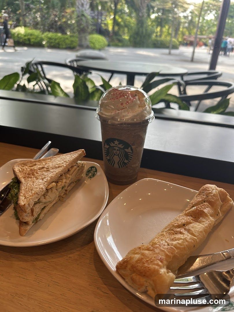 A table at Starbucks with a sandwich, a pastry, and a Frappuccino. The sandwich is on a white plate with the Starbucks logo. The pastry is on another white plate. The Frappuccino is in a clear plastic cup with a dome lid and whipped cream, also with the Starbucks logo visible on the cup. Outside the window, there are tables and chairs, greenery, and people walking.