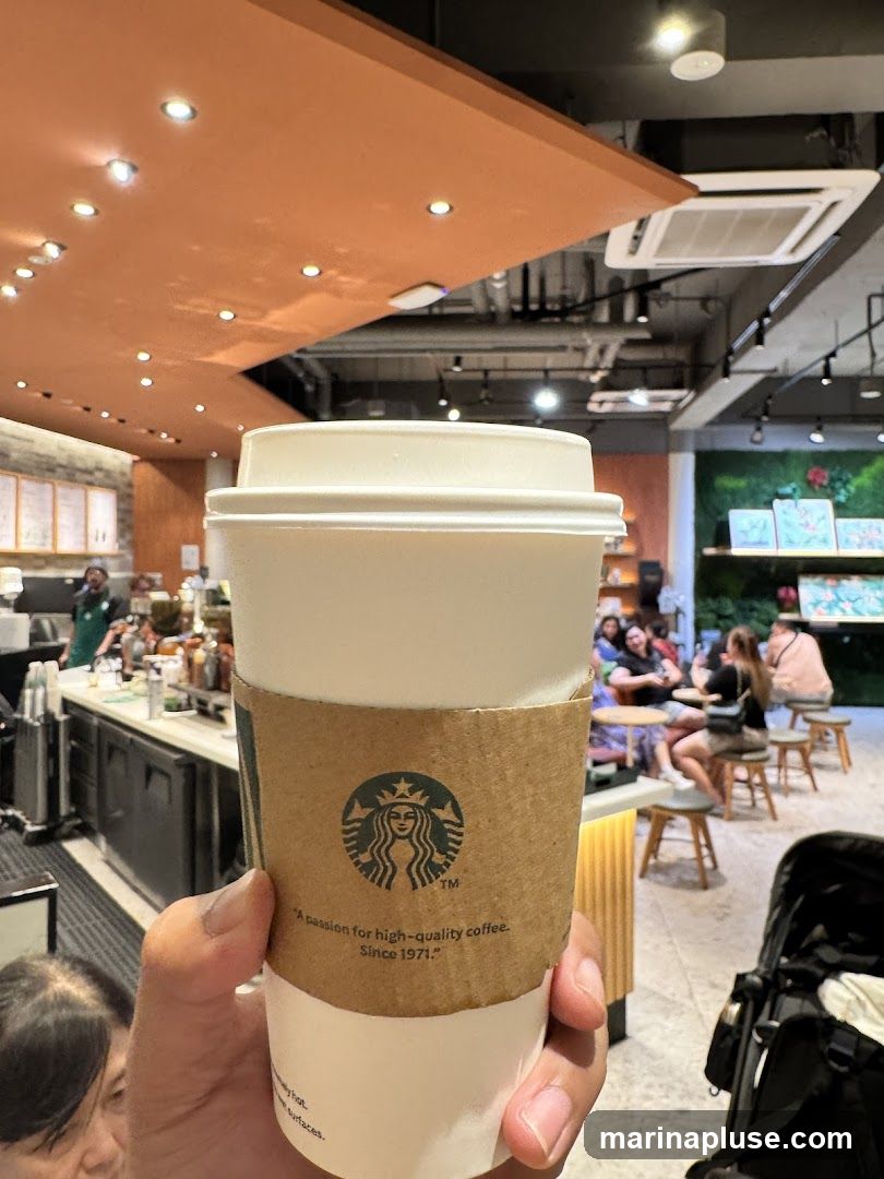 A hand holds a Starbucks coffee cup with a sleeve featuring the Starbucks logo and the tagline "A passion for high-quality coffee. Since 1971." In the background, a cafe interior is visible with customers seated at tables and a counter with baristas.