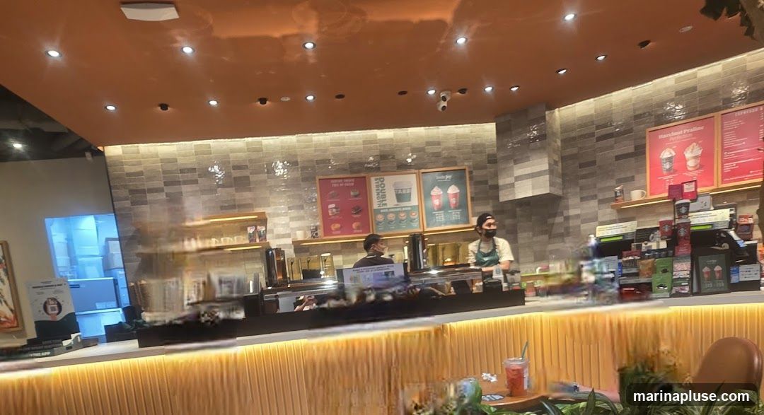 A view of a coffee shop counter with two baristas wearing masks. There are menus displayed on the wall behind them, featuring drinks and food items. The counter is illuminated with warm lighting, and there are various products and merchandise displayed.
