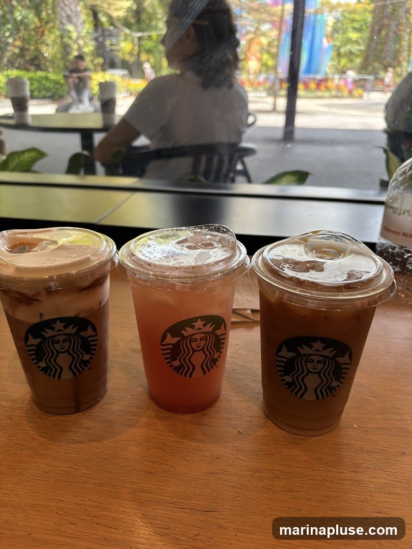 Three Starbucks drinks with the iconic logo are displayed on a wooden table. The drinks appear to be iced coffee, a pink beverage, and another iced coffee. In the background, through a window, people are seated at tables in a bright, outdoor-like setting with greenery and colorful decorations.