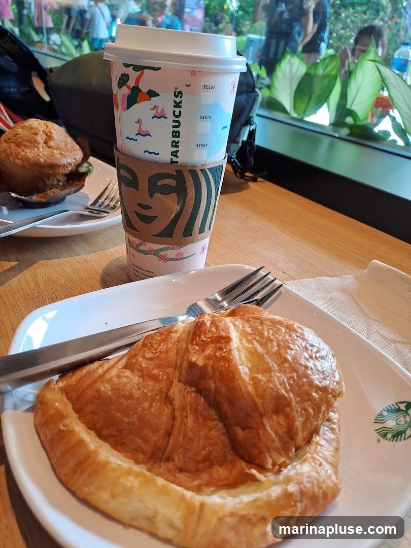 A close-up shot of a Starbucks coffee cup and a croissant on a white plate, with a muffin in the background. The coffee cup has a decorative sleeve with the Starbucks logo and a nature-themed design. The croissant is golden brown and flaky. The scene is set on a wooden table, with a window in the background showing greenery and reflections of people. A fork is placed on the plate with the croissant. The Starbucks logo is also visible on the plate.