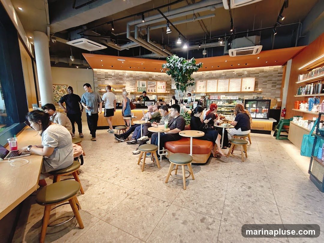 A wide-angle view of a bustling cafe interior. Several customers are seated at tables and a counter, some working on laptops, others chatting. A counter with food displays and menu boards is visible in the background, with staff members attending to customers. The cafe has a modern design with warm lighting and decorative plants.