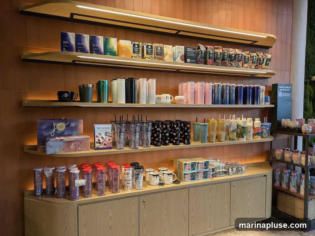 A well-lit display shelf in a Starbucks store showcases a variety of merchandise. The shelves are stocked with packaged teas, coffee bags, tumblers, mugs, and reusable cups. Some items feature festive designs, possibly related to holidays or special collections. A sign on the right reads "COFFEE, SMILE & GIFTS OF JOY".